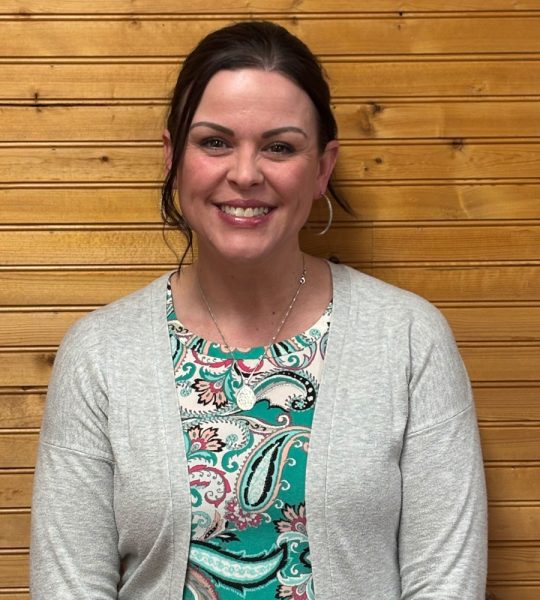 Town of Hartsville Board Member Brinette Steffey wearing a teal, white, black and pink paisley shirt with a grey sweater standing in front of a wood wall