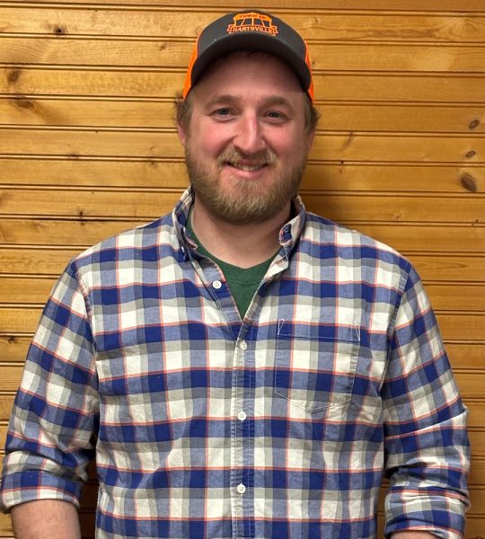 Town of Hartsville Highway Superintendent Jeffrey Getman wearing a blue, white, grey and orange flannel, a ballcap and standing in front of a wood wall